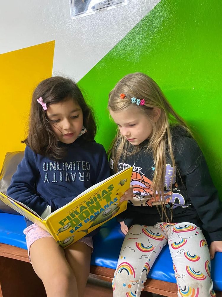 Dos alumnas leen juntas un libro ilustrado en inglés sentadas en una banca